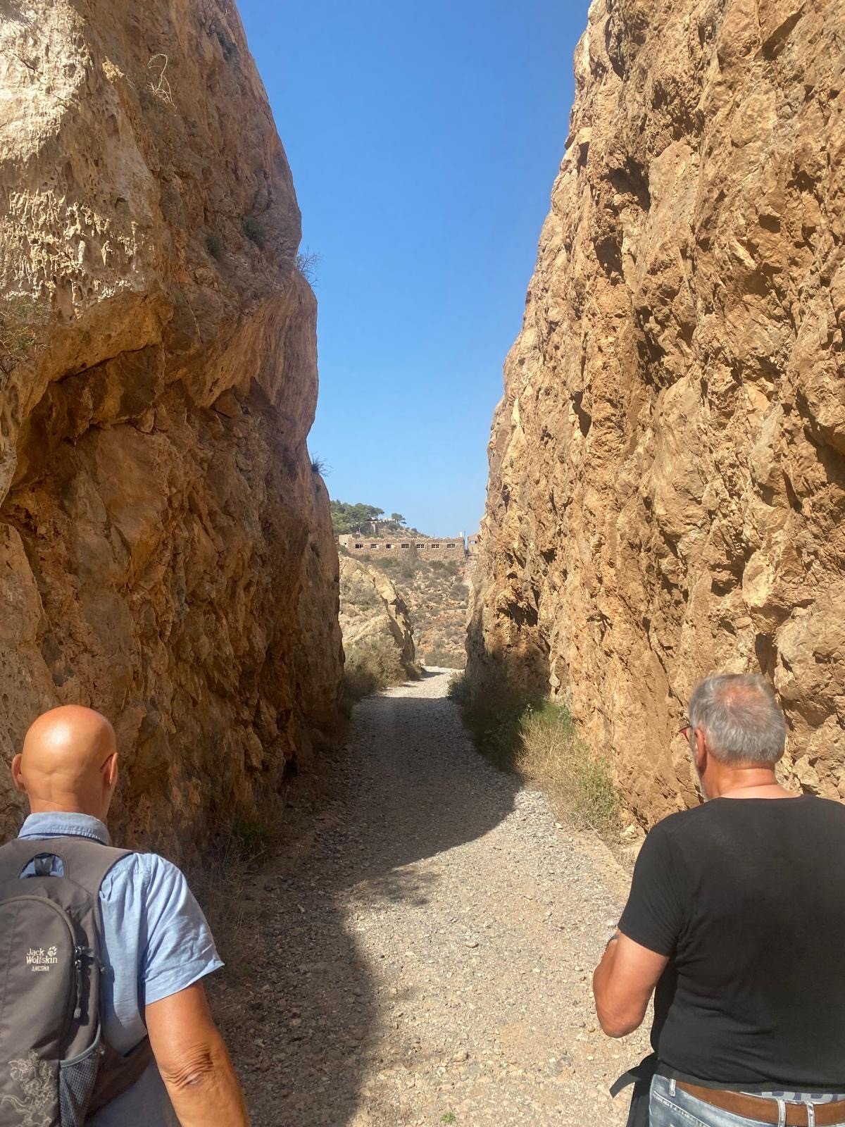 Two walkers passing through a deep cutting with steep ochre rock walls, with the battery on the hill at the far end