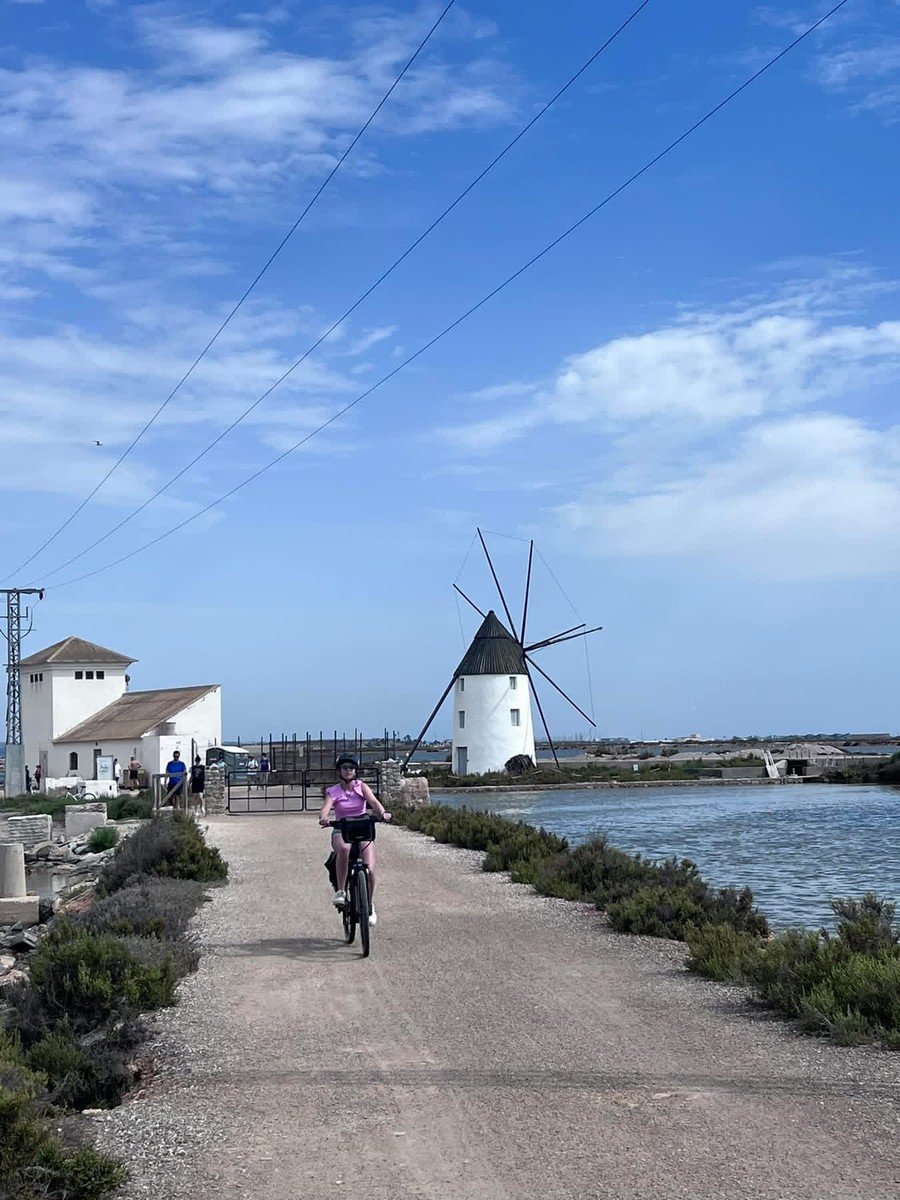 Ciclista en el camino junto a las balsas de sal con un molino salinero hist&oacute;rico al fondo