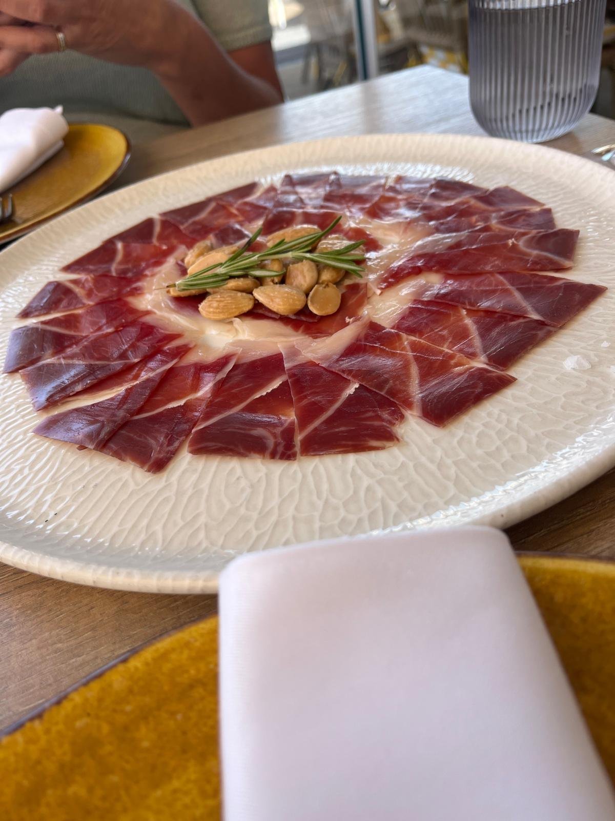 Teller mit d&uuml;nn geschnittenem Ib&eacute;rico-Schinken, Mandeln und Rosmarin auf einer Terrasse am Wasser