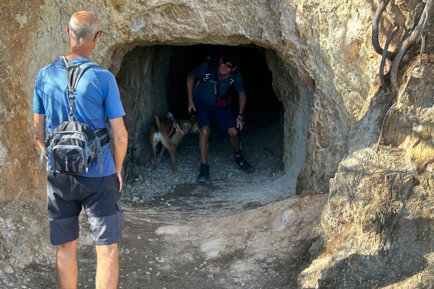 Wanderer mit Hund am Eingang eines in den Fels geschlagenen Tunnels in der Bergbaulandschaft